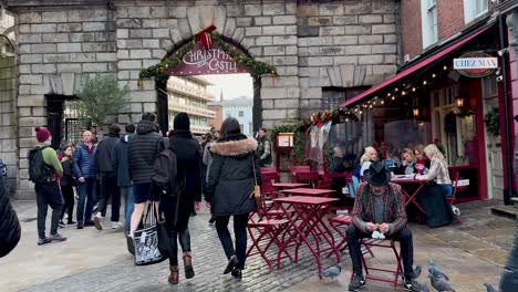 En-4k,-Un-Hombre-Se-Sienta-En-Una-Silla-Mientras-Las-Palomas-Se-Sientan-En-Su-Regazo-Alimentándose-Mientras-Los-Transeúntes-Corren-Hacia-El-Mercado-Navideño-Del-Castillo-De-Dublín-Ignorándolo-Por-Completo.