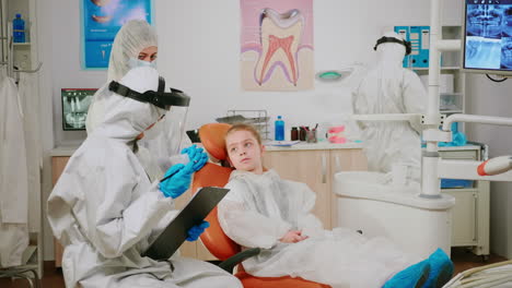 Pediatric-dentist-in-ppe-suit-talking-with-kid-patient-writing-on-clipboard