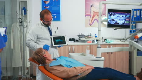 Woman-sitting-on-dental-chair-listening-doctor-looking-on-tablet