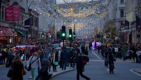 Personas-Con-Bolsas-De-Compras-Cruzan-La-Calle-Entre-Taxis-Negros,-Bicicletas-Y-Rickshaws-Brillantemente-Iluminados-Bajo-Las-Luces-Festivas-En-Regent-Street-Al-Atardecer