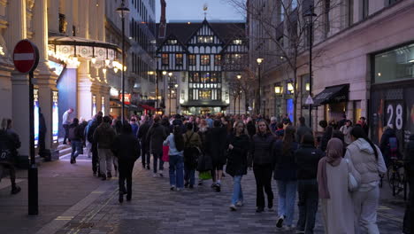 En-Cámara-Lenta,-Multitudes-De-Compradores-Navideños-Caminan-Por-Una-Calle-Lateral-Que-Conduce-Hacia-Los-Grandes-Almacenes-Liberty-Cerca-De-Regent-Street-Al-Atardecer.