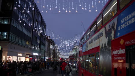 Una-Línea-De-Autobuses-Rojos-En-El-Tráfico-Se-Aleja-Mientras-Los-Compradores-Navideños-Llenan-Las-Aceras-Bajo-Las-Luces-Festivas-En-Oxford-Street-Por-La-Noche