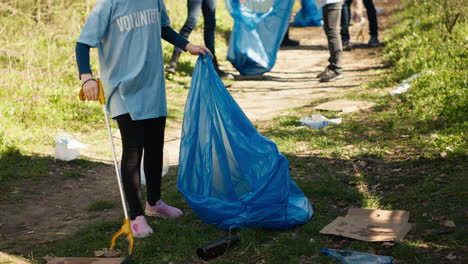 Una-Niña-Pequeña-Recogiendo-Basura-Alrededor-De-Un-Bosque-Con-Una-Herramienta-De-Garra-Y-Bolsas-De-Basura