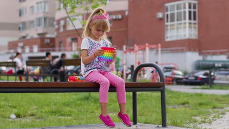 Fröhliches-Mädchen,-Das-Mit-Pop-It-Spielt,-Einem-Einfachen-Sensorischen-Anti-Stress-Spielzeug-Mit-Grübchen-Auf-Einer-Spielplatzbank