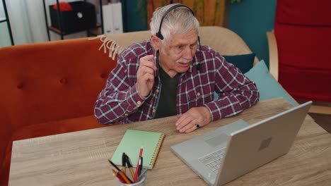 Un-Hombre-Mayor-Independiente-Usa-Auriculares-Para-Hacer-Videollamadas-Hablando,-Enseñando-En-Una-Computadora-Portátil-En-La-Oficina-Del-Hogar