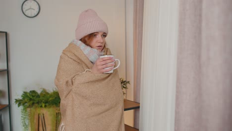 Young-sick-woman-wear-hat-wrapped-in-plaid-stay-alone-shivering-from-cold-on-sofa-drinking-hot-tea