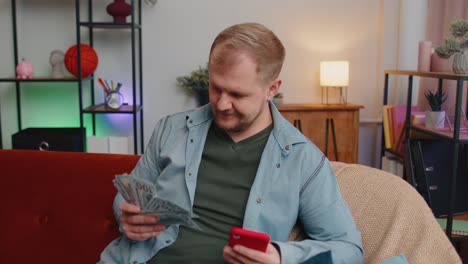 Smiling-happy-young-man-counting-money-cash-and-use-mobile-phone-calculate-domestic-bills-at-home