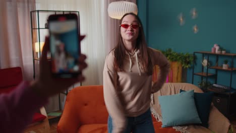 Joyful-blogger-teen-girl-dancing-at-camera-filming-video-using-mobile-phone-at-home-in-living-room