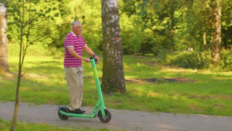 Attractive-active-stylish-senior-man-grandfather-riding-electric-scooter-in-park,-showing-thumbs-up