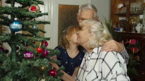 Little-cute-child-girl-with-senior-grandparents-family-decorating-artificial-Christmas-tree-at-home