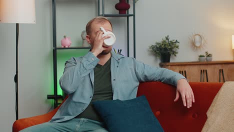 Portrait-of-young-adult-man-sitting-on-couch-relaxing-at-home-room-drinking-enjoy-tea-or-coffee