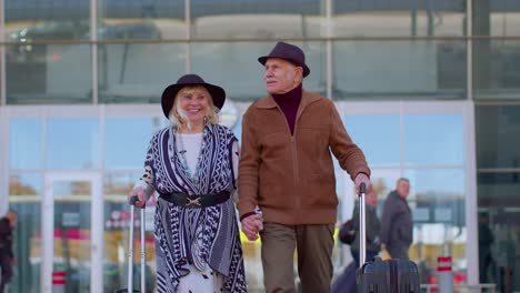 Stylish-retired-family-grandmother-grandfather-walking-with-luggage-suitcase-bags-from-airport-hall