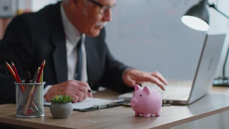 Piggy-bank,-senior-business-man-accountant-typing-on-laptop-computer-calculating-money-income