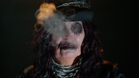 Creepy-mature-woman-in-carnival-costume-of-Halloween-witch-blows-smoke-from-mouth,-trying-to-scare