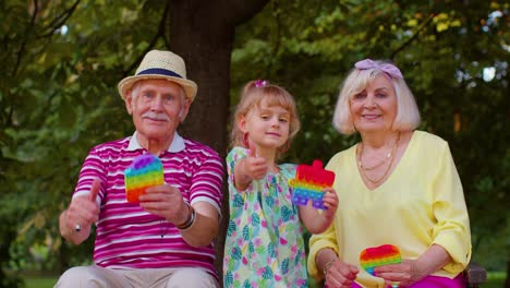 Abuela-Mayor-Abuelo-Con-Nieta-Sosteniendo-Juegos-De-Juguete-Pop-It-Antiestrés-En-El-Parque