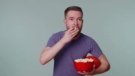 Joven-Barbudo-Aficionado-Al-Baloncesto-Comiendo-Palomitas-De-Maíz-Haciendo-Gesto-Ganador,-Celebrando-La-Victoria