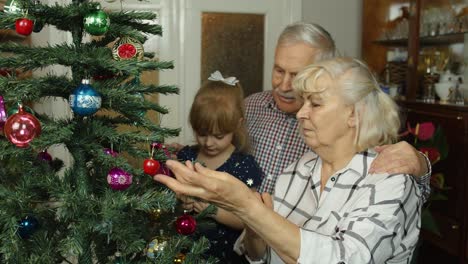 Girl-kid-with-senior-grandma,-grandpa-decorating-artificial-Christmas-tree-with-ornaments-and-toys
