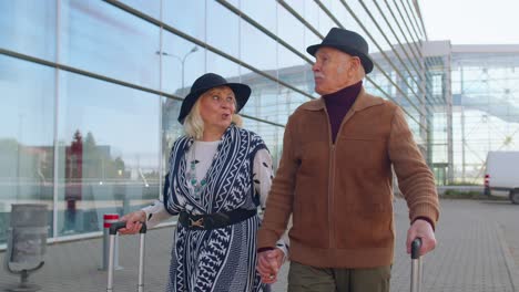 Elderly-husband-wife-retirees-tourists-go-to-airport-terminal-for-boarding-with-luggage-on-wheels