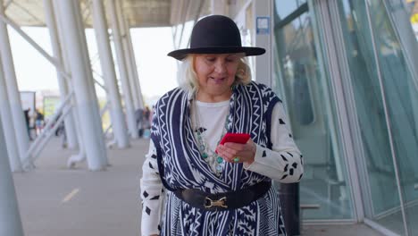 Abuela-Turista-Senior-Caminando-Por-El-Pasillo-Del-Aeropuerto-Internacional,-Usando-Teléfono-Móvil,-Enviando-Mensajes-De-Texto