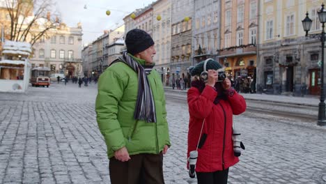 Senior-couple-tourists-grandmother-grandfather-taking-photo-pictures-on-retro-camera-in-winter-city