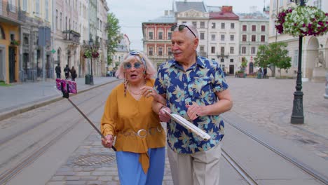 Un-Viejo-Turista-Mayor-Con-Una-Mujer-Caminando-Por-La-Ciudad-Con-Un-Teléfono-Inteligente-En-Un-Palo-Selfie-Teniendo-Una-Videollamada