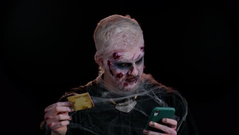 Sinister-man-Halloween-zombie-using-credit-bank-card-and-smartphone-while-purchases-online-shopping