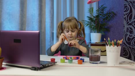 Child-girl-pupil-enthusiastically-drawing-with-brushes-and-paints,-using-laptop-computer-at-home