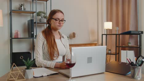Woman-freelancer-drinking-wine-from-glass-after-online-distance-video-call-on-laptop-at-home-office