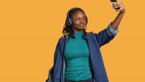 Mujer-Alegre-Con-Auriculares-Usando-El-Teléfono-Para-Tomar-Selfies,-Fondo-De-Estudio