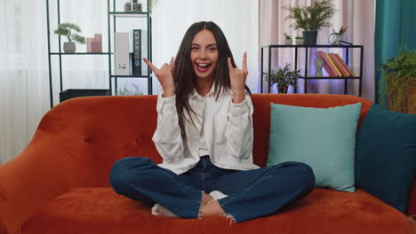 Overjoyed-young-woman-showing-rock-n-roll-gesture,-cool-sign-dancing-rejoicing-in-success-at-home