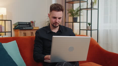 Young-bearded-man-sitting-on-sofa-closing-laptop-pc-after-finishing-business-work-in-room-at-home