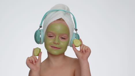 Smiling-child-girl-moisturizing-green-cucumber-mask-on-face-listening-to-music-on-headphones-dancing