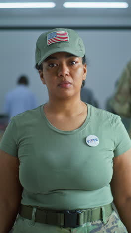 Retrato-De-Una-Mujer-Soldado,-Votante-De-Las-Elecciones-De-Los-Estados-Unidos-De-América.-Una-Mujer-Con-Uniforme-De-Camuflaje-Se-Encuentra-En-El-Colegio-Electoral-Y-Mira-La-Cámara.-Fondo-Con-Cabinas-De-Votación.-Concepto-De-Deber-Cívico.