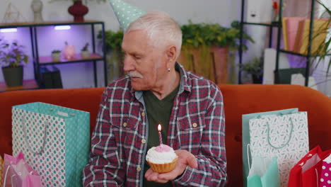 Happy-senior-elderly-grandfather-man-celebrating-birthday-party,-makes-wish-blowing-burning-candle