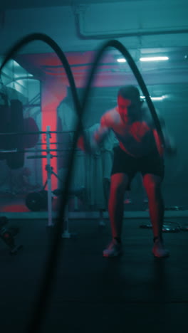 Un-Hombre-Atlético-Entrena-Con-Cuerdas-De-Batalla-En-Un-Gimnasio-De-Boxeo-Oscuro-Con-Iluminación-LED.-El-Boxeador-Masculino-Profesional-Hace-Ejercicios-Cardiovasculares-O-De-Resistencia-Antes-De-Competir.-Tiro-Vertical