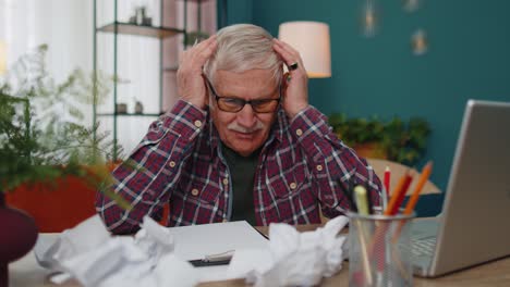 Senior-man-working-on-laptop-computer,-making-notes-on-sheet-of-paper,-upset-by-poor-bad-results