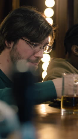 Una-Foto-Vertical-De-Un-Hombre-Con-Gafas-Se-Sienta-En-La-Barra-Del-Bar,-Bebe,-Come-Y-Habla-Con-Un-Amigo-Afroamericano.-Gente-Disfrutando-De-Su-Noche-De-Fin-De-Semana-En-Un-Pub-Deportivo.-Amigos-Pasando-Tiempo-Juntos-En-El-Bar.-De-Cerca.