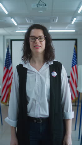 American-female-voter-or-polling-worker-with-badge-walks-and-speaks-on-camera,-calls-for-voting.-National-Election-Day-in-the-United-States.-Voting-booths-at-polling-station.-Civic-duty-and-patriotism