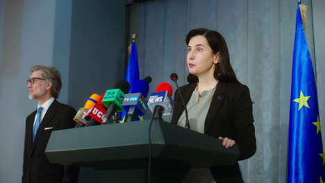 Female-politician-makes-an-announcement,-answers-journalists-questions-and-gives-interview-for-media.-Confident-representative-of-the-European-Union-during-press-conference.-Backdrop-with-EU-flags.