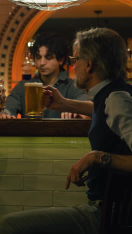 Toma-Vertical-De-Un-Joven-Barman-Da-Vasos-De-Cerveza-A-Amigos-Varones-Maduros.-Dos-Hombres-Hablan-Y-Beben-Cerveza-Sentados-En-La-Barra-Del-Bar-En-Un-Elegante-Pub-Por-La-Noche.-Reunión-De-Viejos-Amigos.-Concepto-De-Estilo-De-Vida-Y-Ocio.