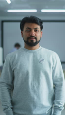 Portrait-of-Indian-man,-United-States-of-America-elections-voter.-Man-stands-in-a-modern-polling-station,-poses-and-looks-at-camera.-Background-with-voting-booths.-Civic-duty-and-patriotism-concept.