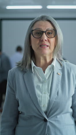 Portrait-of-mature-woman,-United-States-of-America-elections-voter.-Businesswoman-speaks-on-camera-and-calls-for-voting-in-modern-polling-station.-Background-with-voting-booths.-Concept-of-civic-duty.