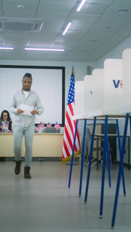 African-American-man-takes-paper-ballot-from-female-polling-officer-at-polling-station-and-walks-to-voting-booth.-National-Election-Day-in-the-United-States-of-America.-Civic-duty-and-democracy.