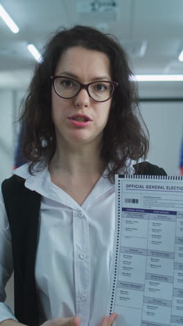 American-woman-speaks-on-camera,-shows-paper-bulletin,-calls-for-voting.-National-Elections-Day-in-the-United-States.-Voting-booths-at-polling-station.-Concept-of-civic-duty-and-patriotism.-Portrait.