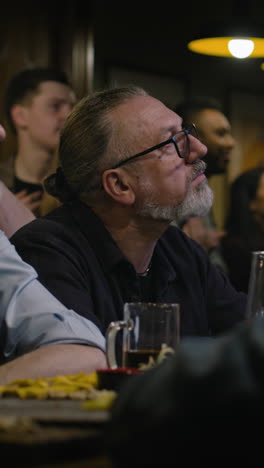 Vertical-shot-of-friends-cheering-for-soccer-team-playing-in-world-league-tournament.-Happy-sport-fans-applaud,-shout-and-drink-beer-watching-football-match-live-broadcast-in-bar-or-sports-pub-at-night.