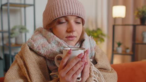 Young-sick-woman-wear-hat-wrapped-in-plaid-sit-alone-shivering-from-cold-on-sofa-drinking-hot-tea