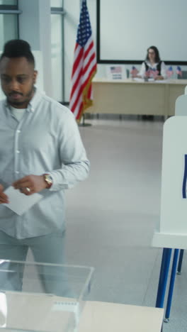 Nervous-African-American-male-voter-chooses-which-presidential-candidate-to-vote-for-in-voting-booth,-looks-at-camera.-US-citizen-at-polling-station-during-Election-Day-in-the-United-States-of-America