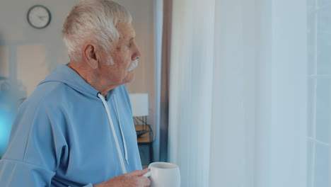 Smiling-senior-old-grandfather-man-drinking-a-cup-of-coffee-or-herbal-tea-at-home-room-near-window