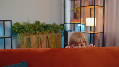 Child-girl-playing-hide-and-seek-peekaboo-game-near-sofa-at-home-alone-looking-at-camera-and-smiling