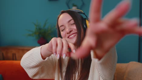 Pretty-Caucasian-young-woman-in-headphones-listening-music-dancing,-singing-in-living-room-at-home